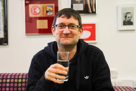 A man with brown hair and glasses sits on a sofa surrounded by pictures. He is wearing a dark hoodie jumper and is holding a glass. The wall behind him is white and covered in large colourful pictures and records.