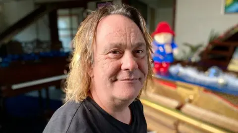 Helen Mulroy/BBC Martin Porter looks directly at the camera and sits in front of the blue upright piano. He has chin length, caramel-coloured hair and is wearing a black T-shirt.