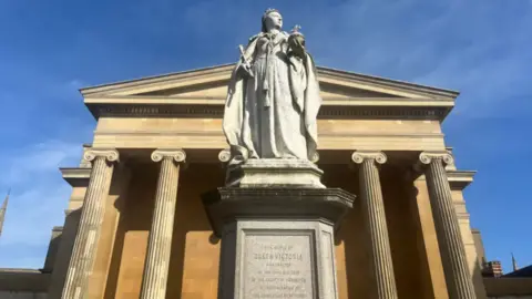 BBC A statue of Queen Victoria holding an orb and sceptre on a plinth outside a sandstone building with tall fluted columns. Blue sky can be seen behind.
