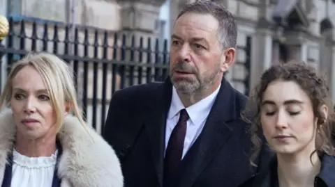 Tony Chambers, with abbreviated  grey hairsbreadth  and a grey beard and wearing a acheronian  bluish  suit, is pictured walking to Liverpool Town Hall flanked by 2  women, 1  blonde and wearing a fur overgarment  and 1  with curly brownish  hairsbreadth  and wearing a acheronian  bluish  jacket