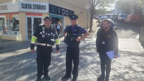 Mike Puckey (left) wearing black trousers and a jacket which has hi-vis yellow bands on the sleeves. The police community support officer Paul Price in the middle wearing a blue short-sleeved shirt with a black police vest and black trousers. On the right is James Mylam from Harbour Housing   who is wearing a black hooded jumper with a black and red lanyard. He is holding a red clipboard in his right hand.