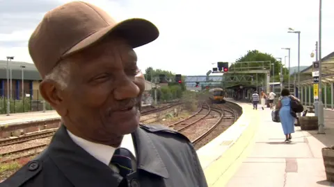 Carlton Green wearing a cap and a shirt and tie. He is looking off camera to his left. There is a railway behind him and a train coming up to the platform.