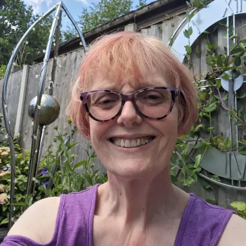Jo Bain Jo Bain wearing glasses and a sleeveless purple top in the foreground, facing the camera in a garden setting. Behind her are flowering plants, climbing greenery, a wooden fence and a large decorative outdoor clock mounted on the fence. A metal garden ornament and blue sky with scattered clouds are visible in the background.