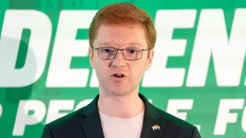 PA Media Ross Greer - a man with red hair, glasses and wearing a dark jacket and white shirt, with Scotland and Pride flag pin badges, speaks while standing in front of a green banner