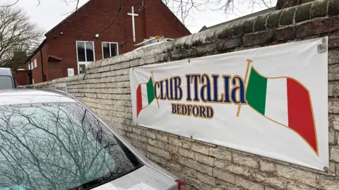 Martin Heath/BBC Banner showing the green, white and red Italian flag and the words "Club Italia Bedford". It is attached to a wall of light brown bricks. There is a church with a white cross on the front wall behind the banner. There is a silver car parked next to the light brown wall.