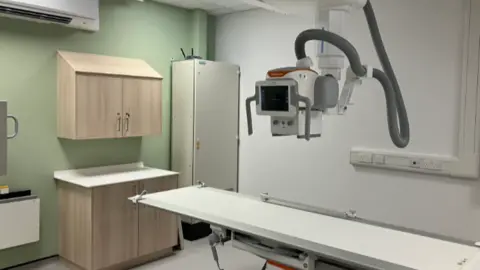 A long white and grey table, with a piece of machinery with a screen at its centre hanging above it. There are cupboards to the left against a light green coloured wall