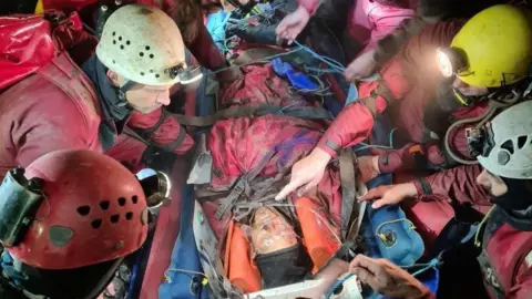 South and Mid Wales Cave Rescue George Linnane is underground lying on a stretcher. He is surrounded by a rescue team who are wearing safety helmets with torches on the front of them.