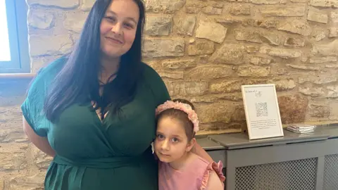 Lucy Bateman a woman dressed in a dark green wraparound playsuit with her arm around a little girl in a pink dress with a pink flowery headband