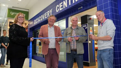 Mikal Ludlow A woman and three men, all smiling to camera, and holding a blue ribbon, standing in a shopping centre outside a store called 'Discovery Centre'