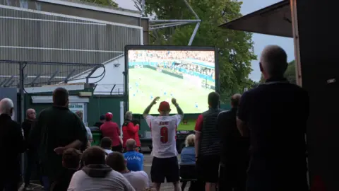 Dan Howell Football fans watching a match on a big screen at Yeovil Town Football Club. There is a man in the centre of the frame wearing a white England top with Kane 9 on the back. His arms are up in the air.