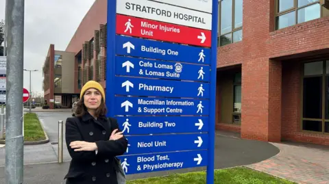 Manuela Perteghella A woman wearing a black coat and yellow beanie hat stands in front of the Stratford Hospital sign. The sign includes directions and names of the various hospital departments and units.