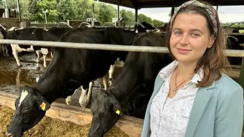 BBC A woman with brown hair and a floral headband is standing in front of a cattle enclosure on a farm. There are black and white cows putting their heads through a fence and eating hay off the ground.