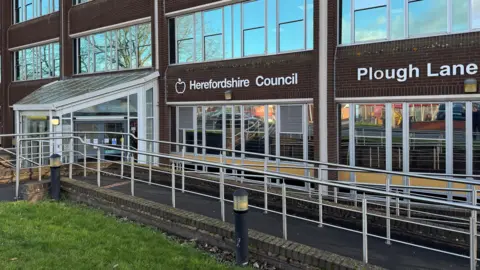 The main entrance of a brick building, with a large white logo featuring the words 'Herefordshire Council'.