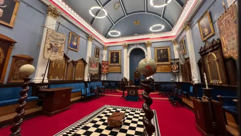 It's a wide shot of the inside of the Freemason Temple. The carpet is dark red and there's a black and white squared section in the middle. There are wooden stands on the edge of the carpet. There are two small globes on wooden stands. Decorated flags marking the lodges of Freemasons in Jersey are displayed around the room. The walls and ceiling is painted light blue.