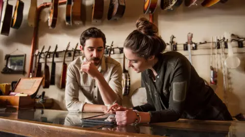 A young couple, a man and a woman, standing behind a wood and glass counter in a music shop. They are looking down at tablet device presumably displaying information about their business. Two racks of guitars are hanging behind them.