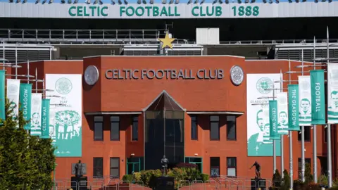 PA Media The front of Celtic's stadium. It is in red brick with green writing and the club emblem on it. There are green and white banners leading up to the entrance with former players and managers' faces on them.