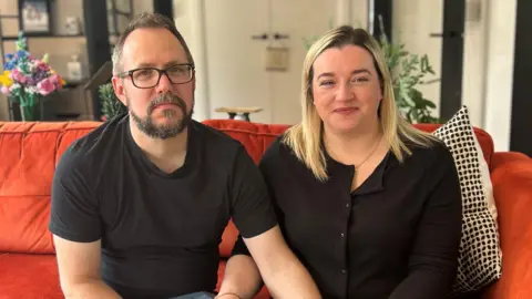 A man and woman are looking directly at the camera. He has dark hair and glasses and is wearing a black t-shirt. The woman has blond hair and is wearing an all black outfit. The pair are sitting on an orange sofa.