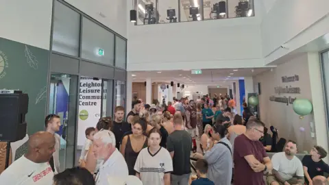 Everyone Active The inside of the centre, with crowds of people inside the corridor. Above the corridor you can see windows onto the gym, with fitness machines. 