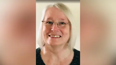 Stella Parkes Stella in a professional close up shot, smiling to camera. She has frameless glasses and white hair down to her shoulders and is wearing a black top. There is an off-white background behind her