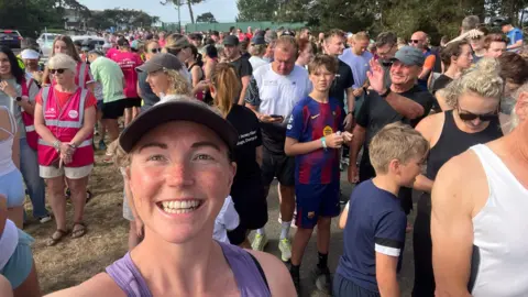 Runner Alice Harvey, who is wearing a baseball cap and a purple vest. Takes a selfie at the start line of Jersey Parkun. Hundreds or people of all ages are stood behind her. Harvey is smiling.