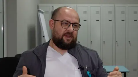 Un hombre calvo con barba y gafas se sienta en una habitación con taquillas al fondo.