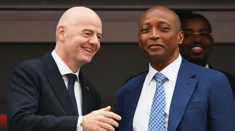 Fifa president Gianni Infantino, wearing a black jacket, white shirt and dark blue tie, smiles and holds up his right arm as he speaks to Caf president Patrice Motsepe, who is wearing a blue suit jacket, white shirt and blue patterned tie. The pair stand close together and are visible from the stomach up