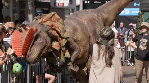 The photo shows a dilophosaurus puppet. It has a brown body, and an orange spike on its head. There are crowds of people behind a fence watching it parade around.