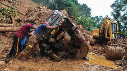 Getty Images Rescue workers push an overturned vehicle stuck in the mud and debris at a site of a landslide said to have been caused by heavy rains in Kokkayar in India's Kerala state, 17 October 2021