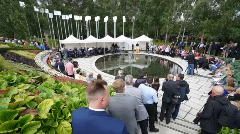 PA Media A group of people gather in the memorial gardens in Omagh, which features a small pond and 29 pillars to represent the victims, for the 25th anniversary of the attack