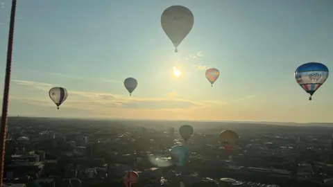 Several hot air balloons flying over Bristol while the sun rises. They are all different shapes and sizes.