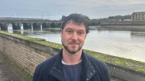 BBC A man wearing a blue jumper and blue coat with dark brown wavy hair standing in front of a river