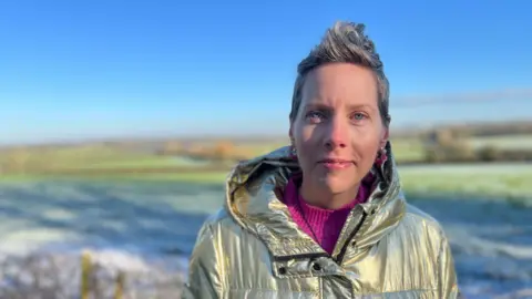 Laura Rawlings pictured outside, in front of frosty fields and blue sky, blurred in the background. She is wearing a gold metallic puffer jacket, a bright pink jumper, large earrings and has short grey hair. 