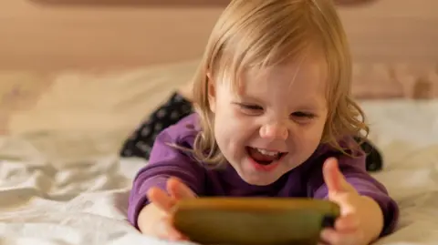 Getty Images Girl with blonde hair aged about three is lying on her stomach on a bed with white bedsheets. She is holding a phone in her two hands and is smiling with her nose scrunched. She looks happy.