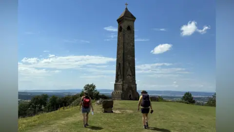 BBC/Steve Mellen A tall narrow brick tower rises up into a blue sky at the top of a hill. There are two hikers with backpacks on their backs and sun hats walking towards the tower.