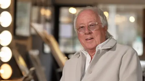 Piers Hopkirk/BBC Brian Aris pictured in a photography studio. Brian, who is wearing a pale grey jacket and a white shirt, has white hair and glasses.