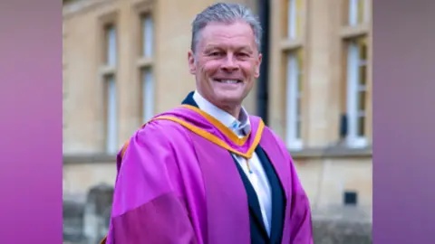 Steve Cotterill, manager of Cheltenham Town FC, smiles as he stands in purple robes outside Cheltenham Town Hall. He is wearing a white shirt and suit jacket underneath.