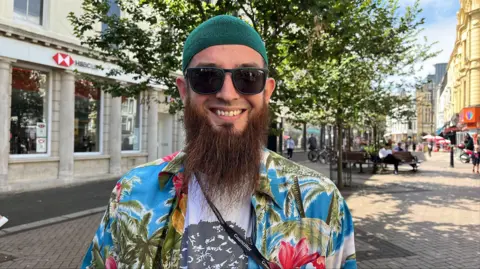CHRISSIE REIDY/BBC Yunis Smith, a man with sunglasses and a long beard smiles at the camera