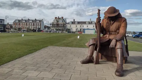 Colin Park/Geograph The Tommy statue shows a giant World War One soldier sat on a trunk. He is holding a gun. It is made out of brown metal and is next to the the green.