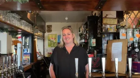 Morgan Webb/BBC Woman with a black shirt and blonde hair smiling into the camera, behind a pub bar with beer taps. 