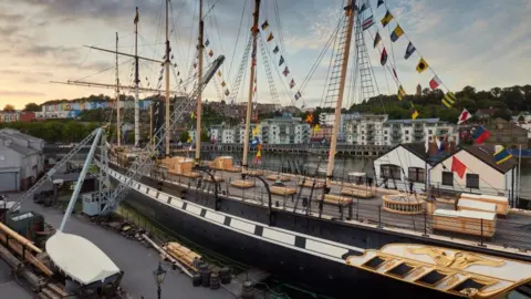 The SS Great Britain, a giant black ship in Bristol Harbour, lined with coloured flags. There are rows of colourful terraced homes in the background.