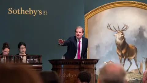 Rayan Bamhayan, Sotheby's Auctioneer with his hand raised in front of the Scene of Braemar painting. Sothebys is written on the wall behind him