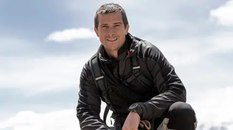 Bear Grylls crouching down, wearing a black jacket and mountain gear. Mountain tops are just visible behind him.