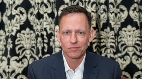 Nordin Catic/Getty Images para The Cambridge Union Peter Thiel con una camisa blanca y una chaqueta azul frente a una pared floral
