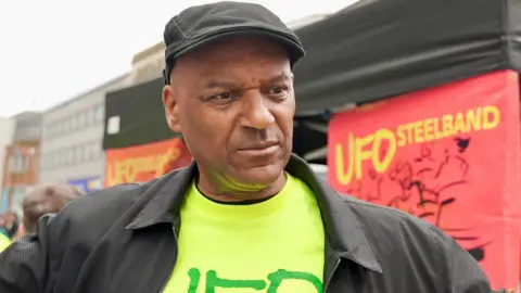 Colin Salmon is wearing a flat cap and a bright green top and stood in front of a red banner that says "UFO STEELBAND" in yellow text.