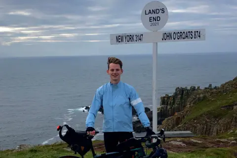 Tom Williamson Tom Williamson in a blue jacket holding up a bicycle and smiling for the camera. He is next to a sign that reads Land's End and points towards New York with a distance of 3147 listed and towards John O'Groats with 874 listed. The sea is visible behind him.