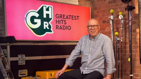 Greatest Hits Radio Ken Bruce in front of the Greatest Hits Radio logo