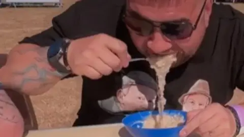 A man spoons porridge into this mouth. It is spilling into a plastic blue bowl. He is wearing sunglasses.