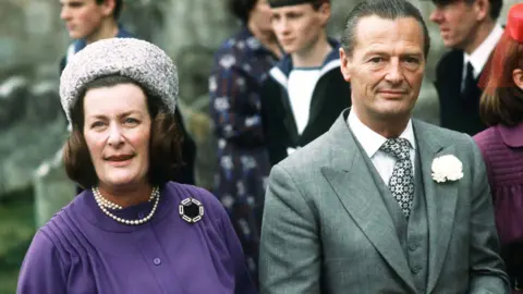 Getty Images Pamela and David Hicks. She is wearing a purple dress with a grey hat, and he wears a grey suit with a white flower attached. 
