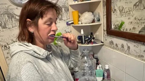 A woman brushing her teeth while holding a bottle of water in her bathroom in a house in Bourton-on-the-Water