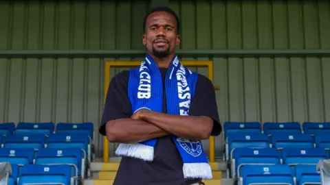 Manny Duku, wearing a blue Eastleigh scarf, poses for a photo at the club's stadium